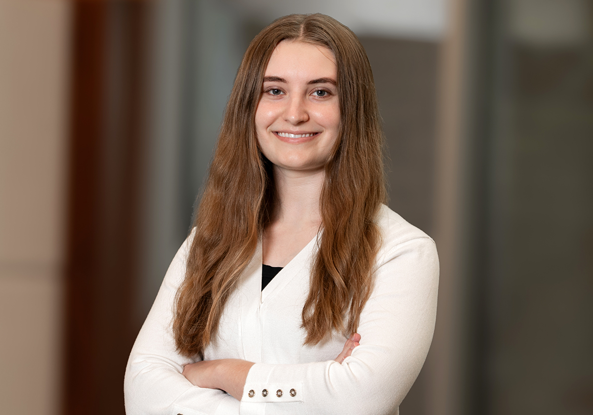 Professional portrait of a young woman with long light brown hair, arms crossed, wearing a white blazer and smiling in a neutral office setting.