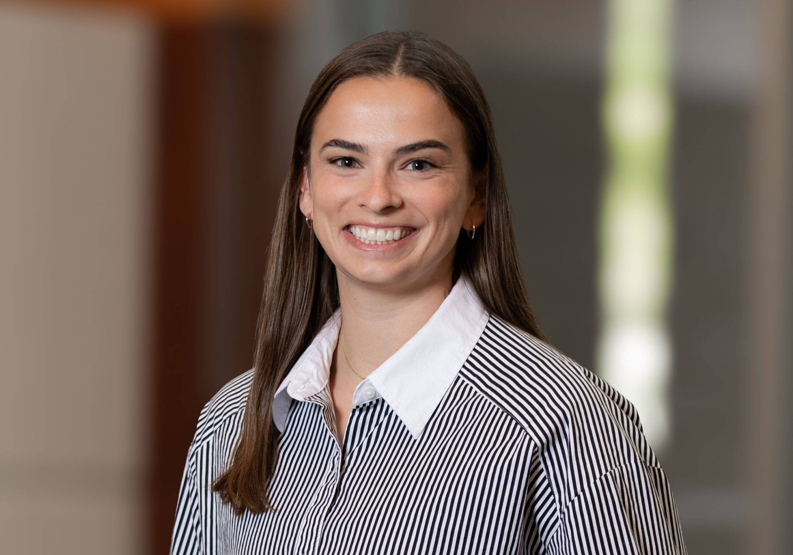 Portrait of a smiling woman with long brown hair, wearing a white collared shirt with black stripes. (Indoor backdrop)
