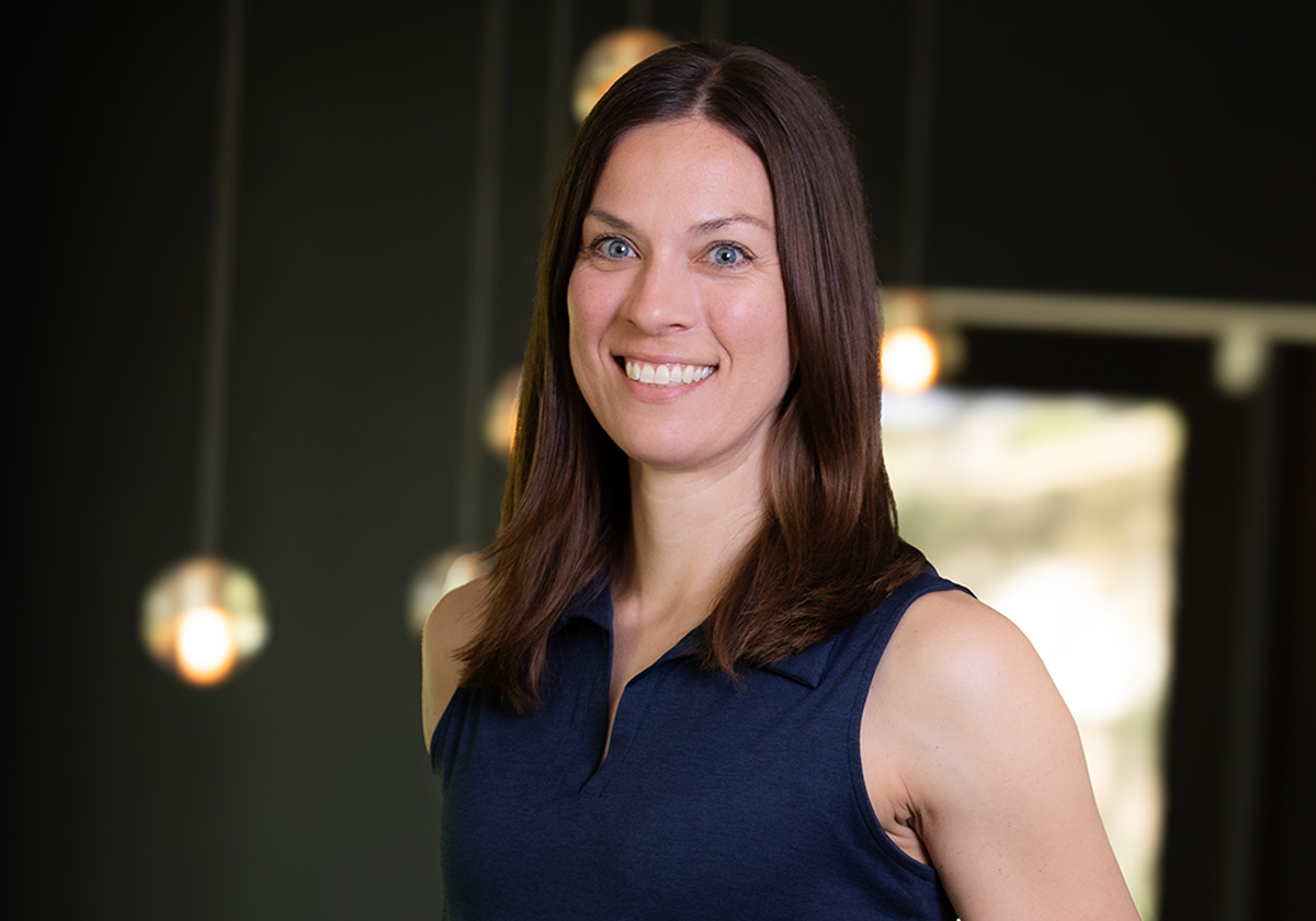 Portrait of a smiling woman with shoulder-length brown hair wearing a navy sleeveless top, in a modern indoor space with blurred lights in the background.