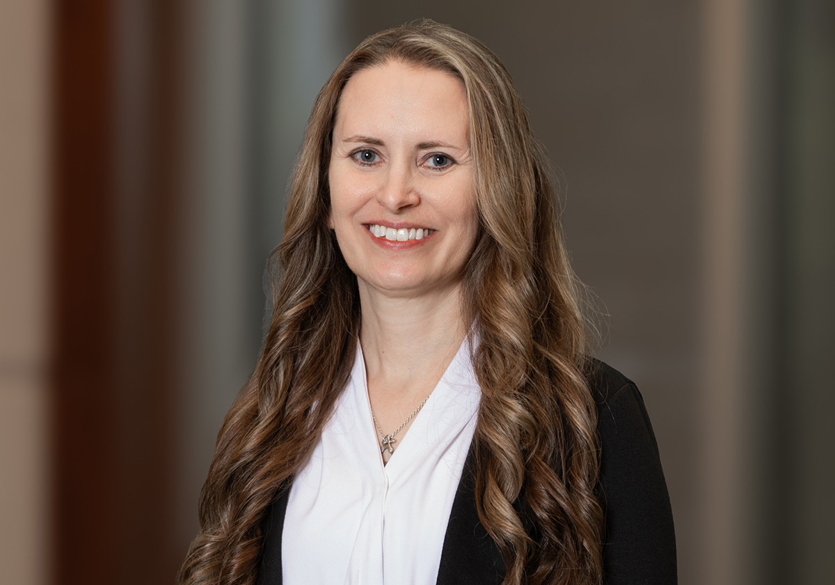 Professional headshot of a smiling woman with long, wavy light brown hair, wearing a white blouse and black blazer.