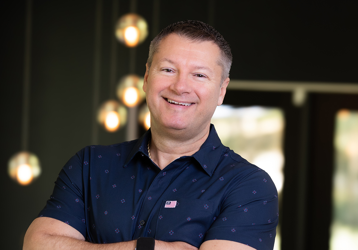 Smiling man in a navy patterned shirt, arms crossed, in a modern office setting with warm pendant lights in the background.